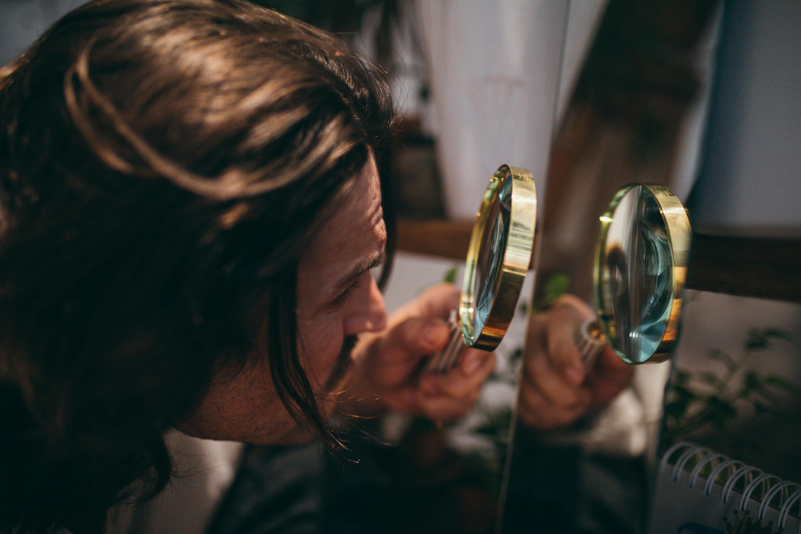 Close-up shot of a man with long hair using a magnifying glass to inspect his reflection in a mirror.